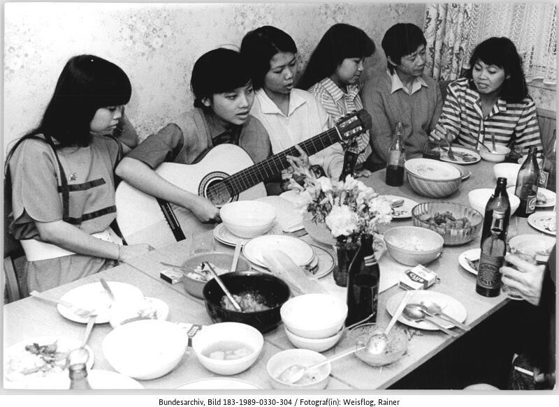 Freizeit vietnam. Gastarbeiter, Cottbus (1989). Im Original: „Nguyen Thi Hong Hou feiert im Wohnheim ihren [20.] Geburtstag. Nach dem Essen werden Volks- und Liebeslieder der Heimat gesungen, (…).“ (c) BArch, Bild 183–1989–0330–304, Foto: R. Weisflog